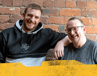 Artwork for the NHS West Yorkshire ICB: Check in with a Mate campaign featuring a photograph of two smiling men standing together against a brick wall with an overlaid painted texture.