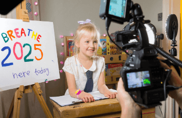 A photograph of a young girl in front of a film set up. She stands next to an easel with the words breathe 2025 here today in colourful, hand drawn letters. Part of the Wakefield Council: Breathe 2030 project.
