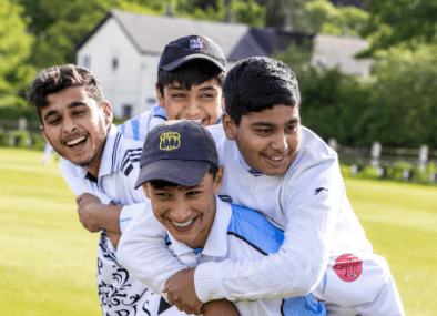 A photograph of four young boys in cricket whites, hugging and laughing on a cricket field from the Active Bradford: Join Us: Move Play campaign