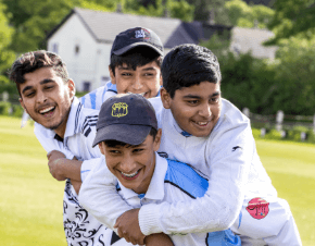 A photograph of four young boys in cricket whites, hugging and laughing on a cricket field from the Active Bradford: Join Us: Move Play campaign