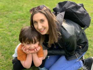 Helena Hamilton kneeling on the grass next to her 3 year old son. She has sunglasses on her head and a backpack on. Her son is cradling his chin in his hands