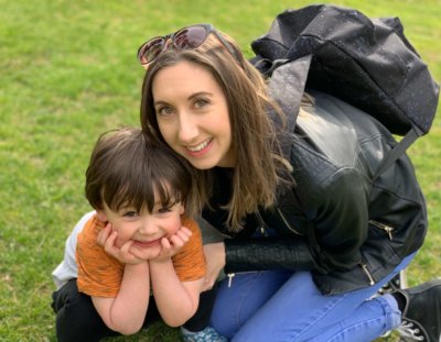 Helena Hamilton kneeling on the grass next to her two year old son. She has sunglasses on her head and a backpack on. Her son is cradling his chin in his hands