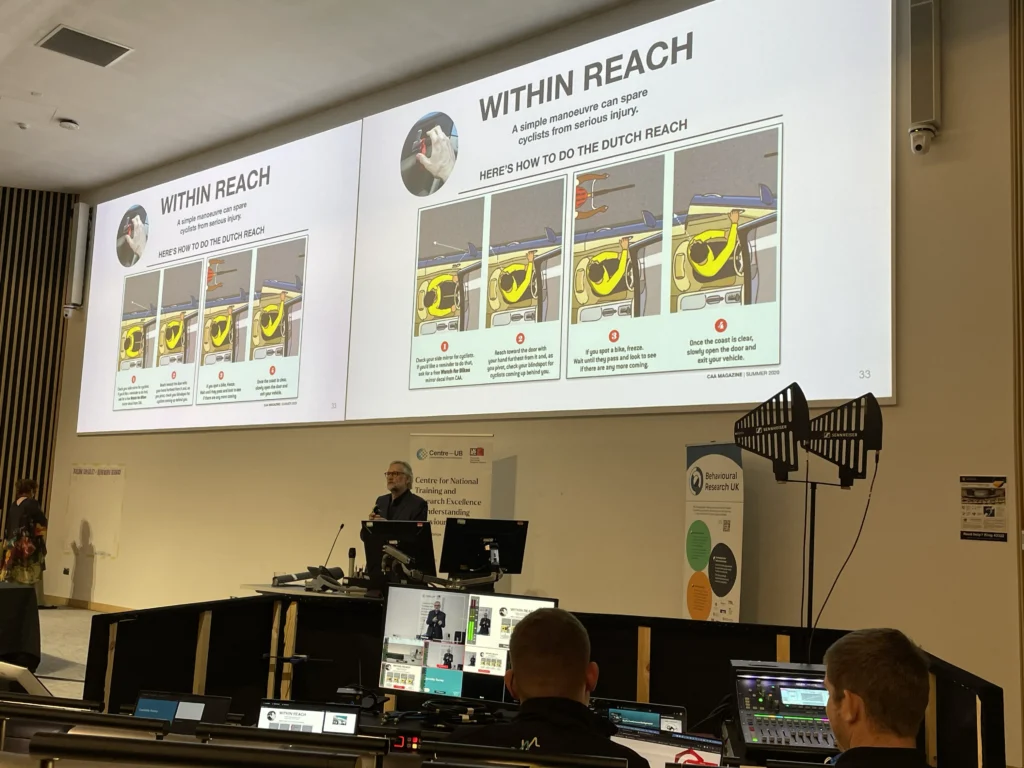 Large screen presentation in an auditorium stating 'within reach, Here's how to do the dutch reach.'